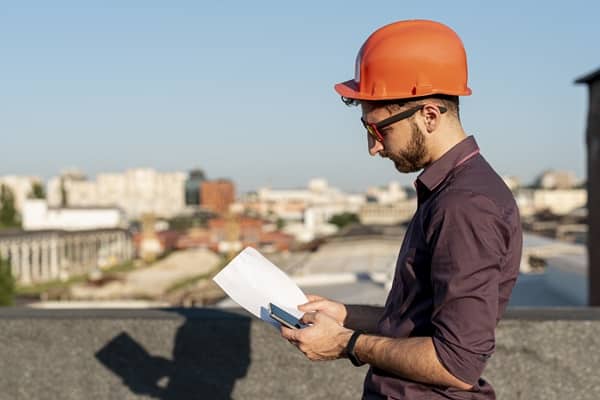 Homme portant un casque de chantier orange, consultant des plans et un téléphone sur un toit avec une vue panoramique sur une zone urbaine, représentant le travail du géomètre-expert.